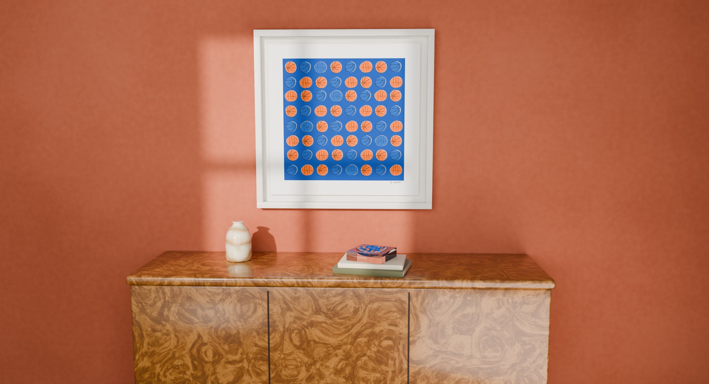The Champions Edition candy dish on two art books on a burl-wood sideboard, under Robyn's framed Champions Edition print, against a terracotta wall